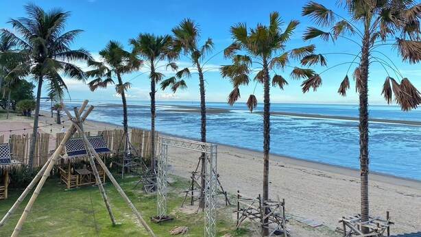 Uma praia com palmeiras, uma cerca de madeira e uma estrutura com telhado de palha.