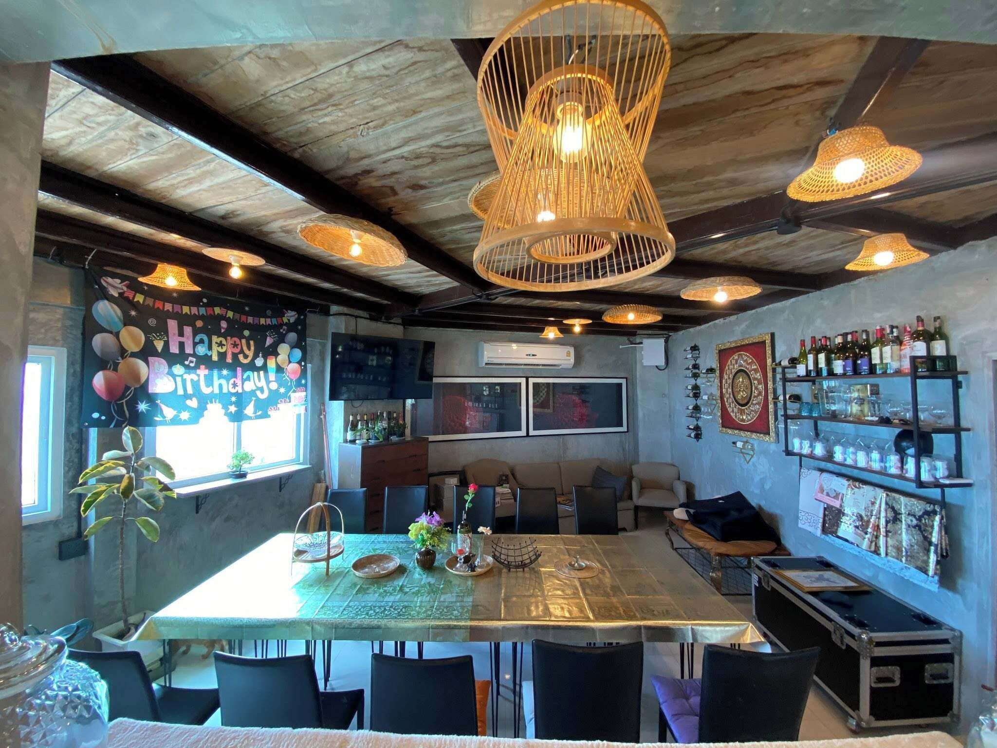 A restaurant with a high ceiling, exposed beams, and a variety of hanging lights. There is a bar area with shelves of bottles and glasses, and a table set for dining.