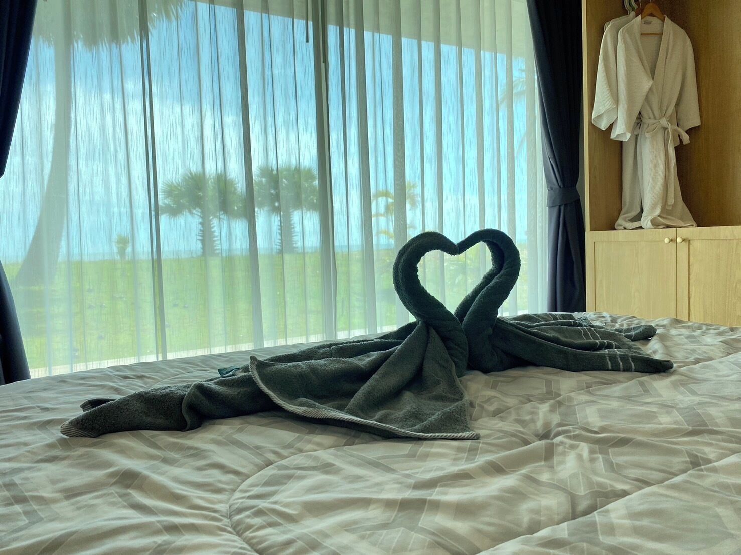 A bed with a heart-shaped towel arrangement and a robe hanging on a wooden cabinet.