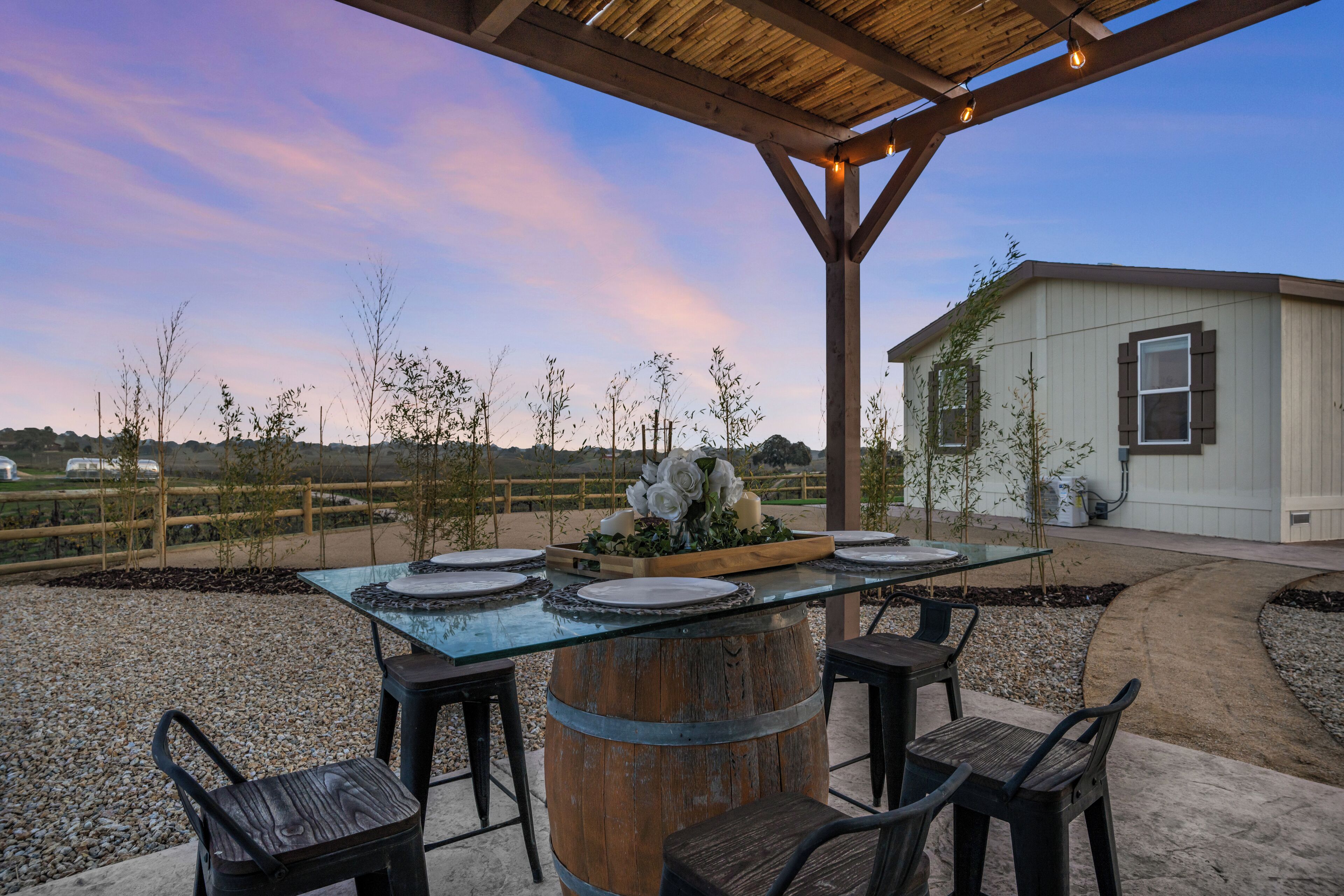 An outdoor patio with a glass-top table surrounded by chairs, a wooden barrel centerpiece, and a small building in the background.