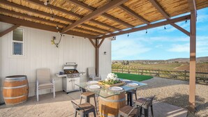 Un patio al aire libre con una mesa de vidrio rodeada de taburetes de madera, una parrilla y un barril rústico.
