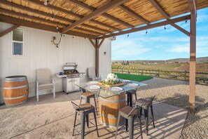 An outdoor patio with a glass-top table surrounded by wooden chairs, a barbecue grill, and a rustic barrel. - Near Tasting Rooms + Outdoor Dining + Views | Sip and Stay at Vahali Vineyards (Paso Robles)
