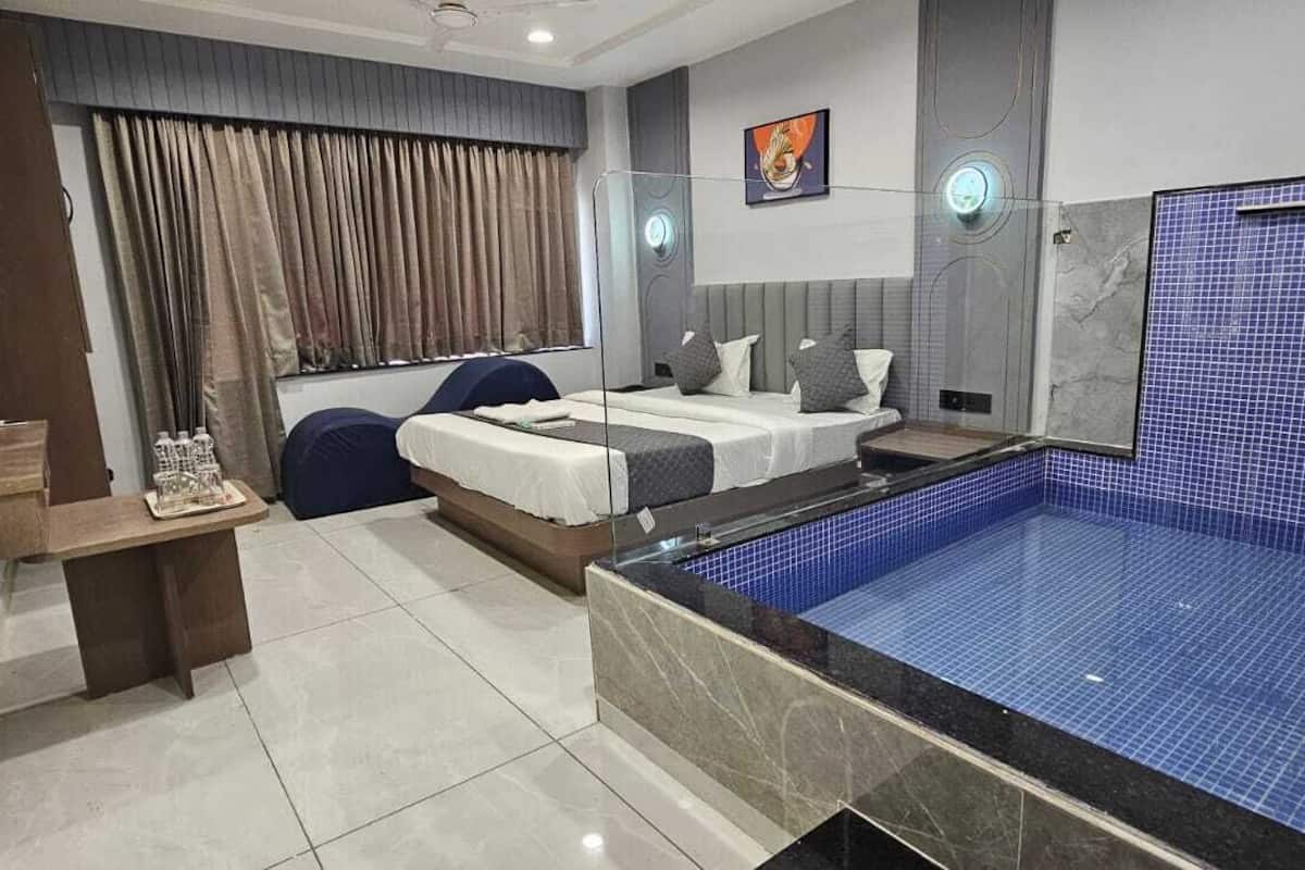 Une chambre d’hôtel moderne avec une grande piscine aux carreaux bleus, un lit avec une tête de lit, une table de chevet et une fenêtre avec des rideaux.