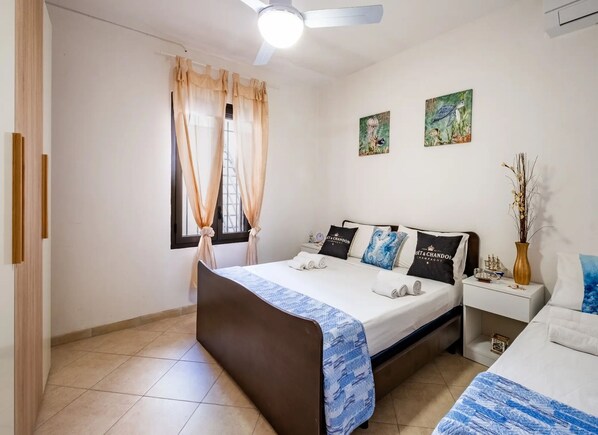 Un dormitorio con cama, mesita de noche, ventana con cortinas, ventilador de techo y dos cuadros colgados en la pared.