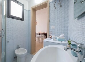 Un baño con bañera, lavamanos y bidé. Hay una ventana sobre el lavamanos y una puerta que da a un comedor.