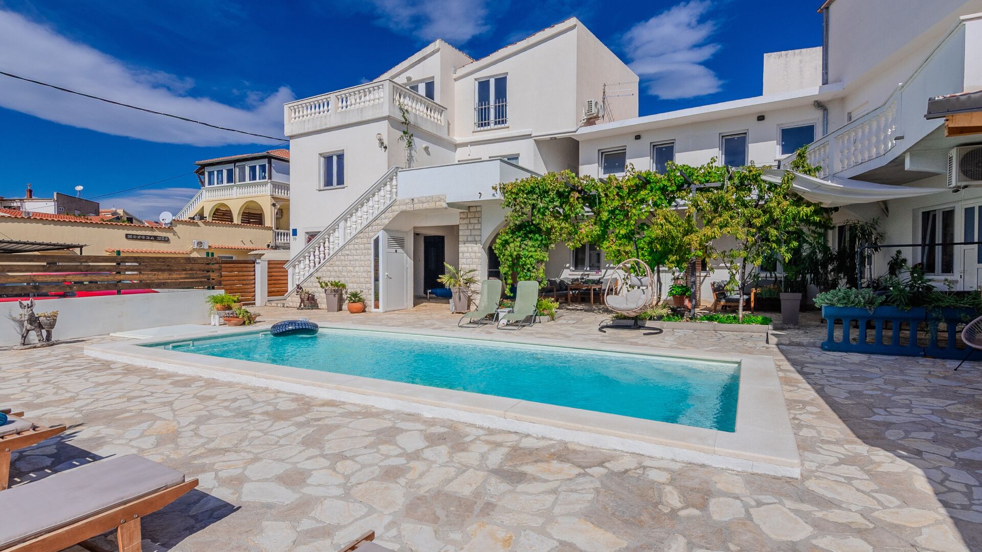 A poolside area with a stone floor, a pool, lounge chairs, and a staircase leading to a second floor.