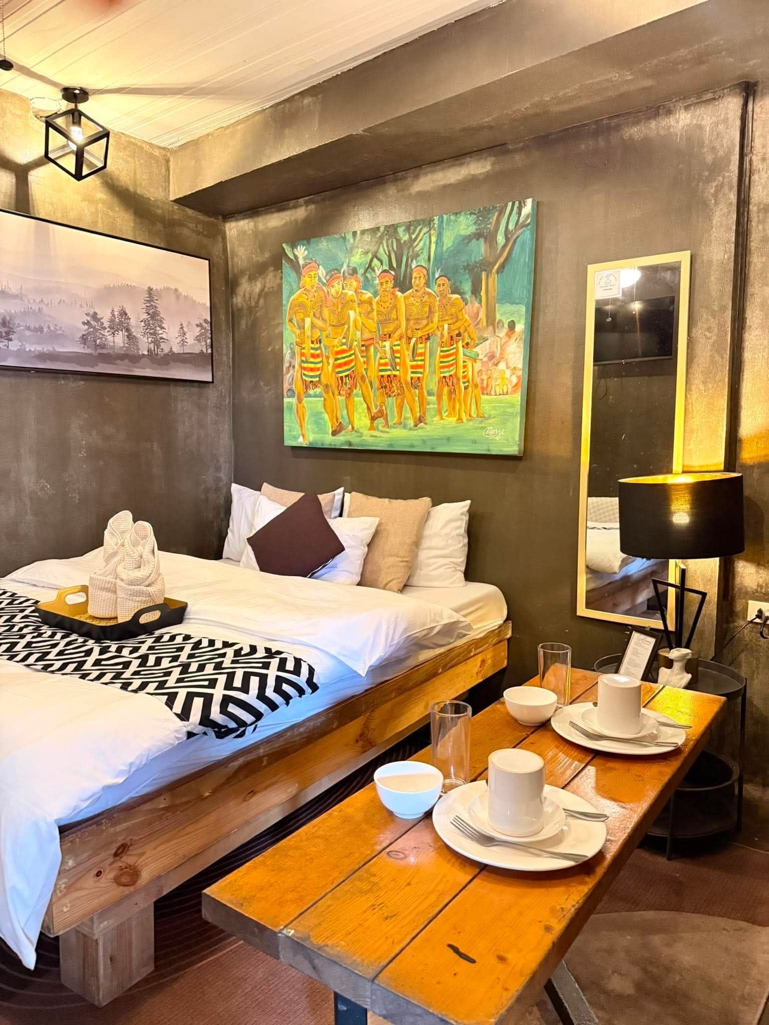 A hotel room with a bed, a painting, a table set for breakfast, and a lamp.