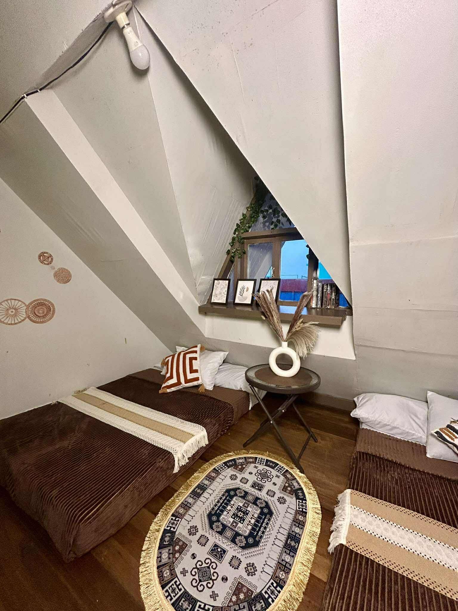 A compact, triangular room with a sloped ceiling, a small round table, and two beds with patterned bedding.