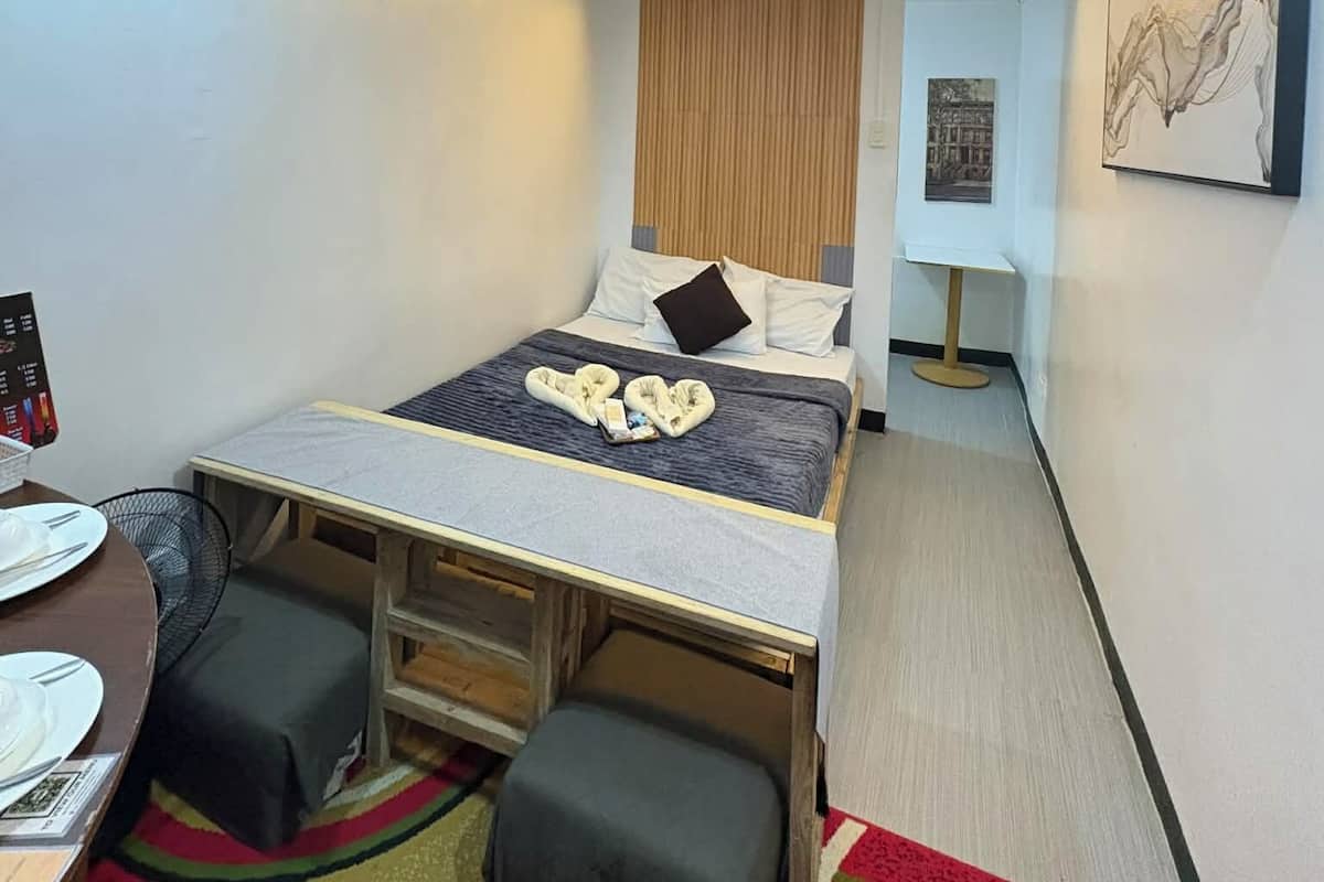 A compact hotel room with a bed, a small table, a chair, a bench, and a wall-mounted artwork.