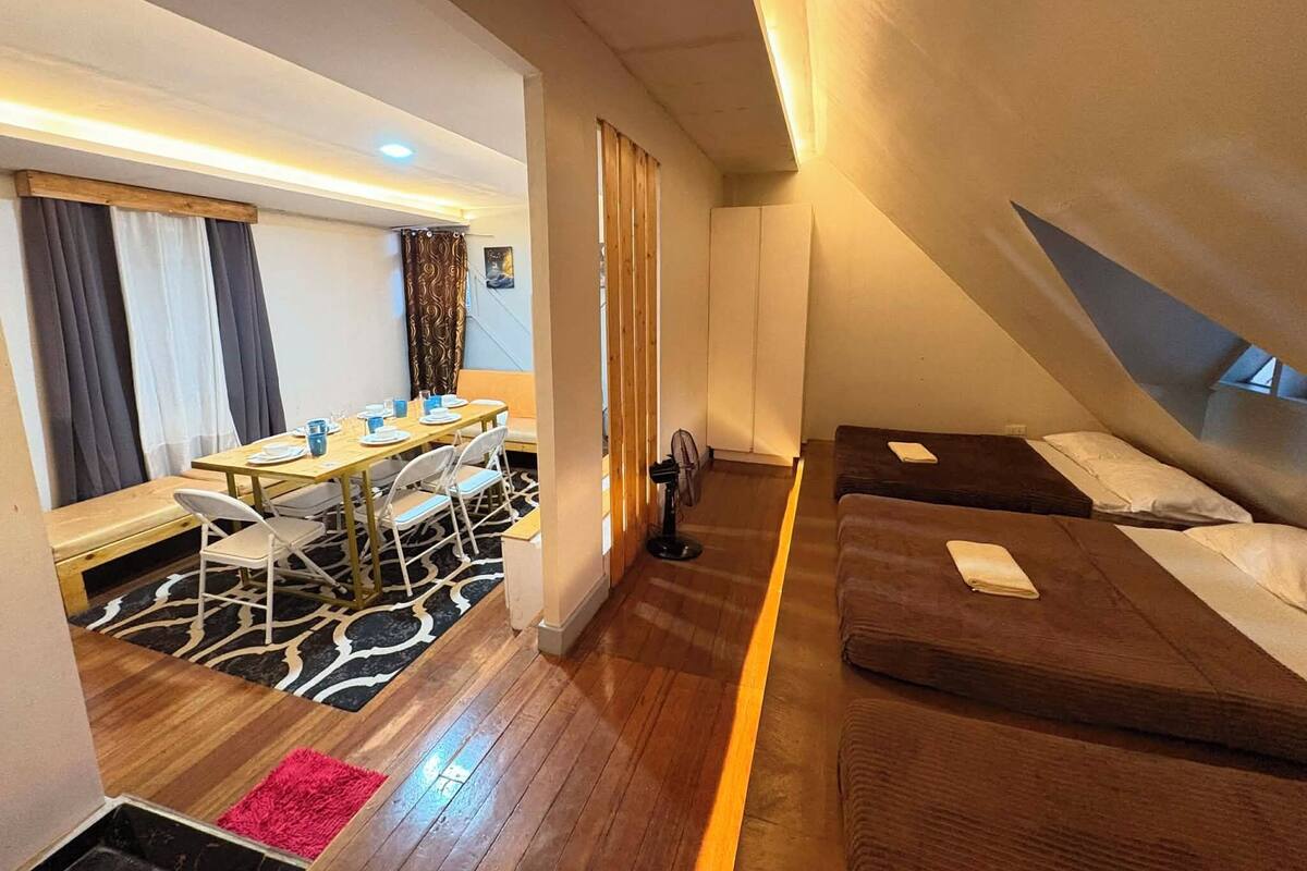 A compact hotel room with a dining area, two single beds, a small sitting area, and a ceiling fan.