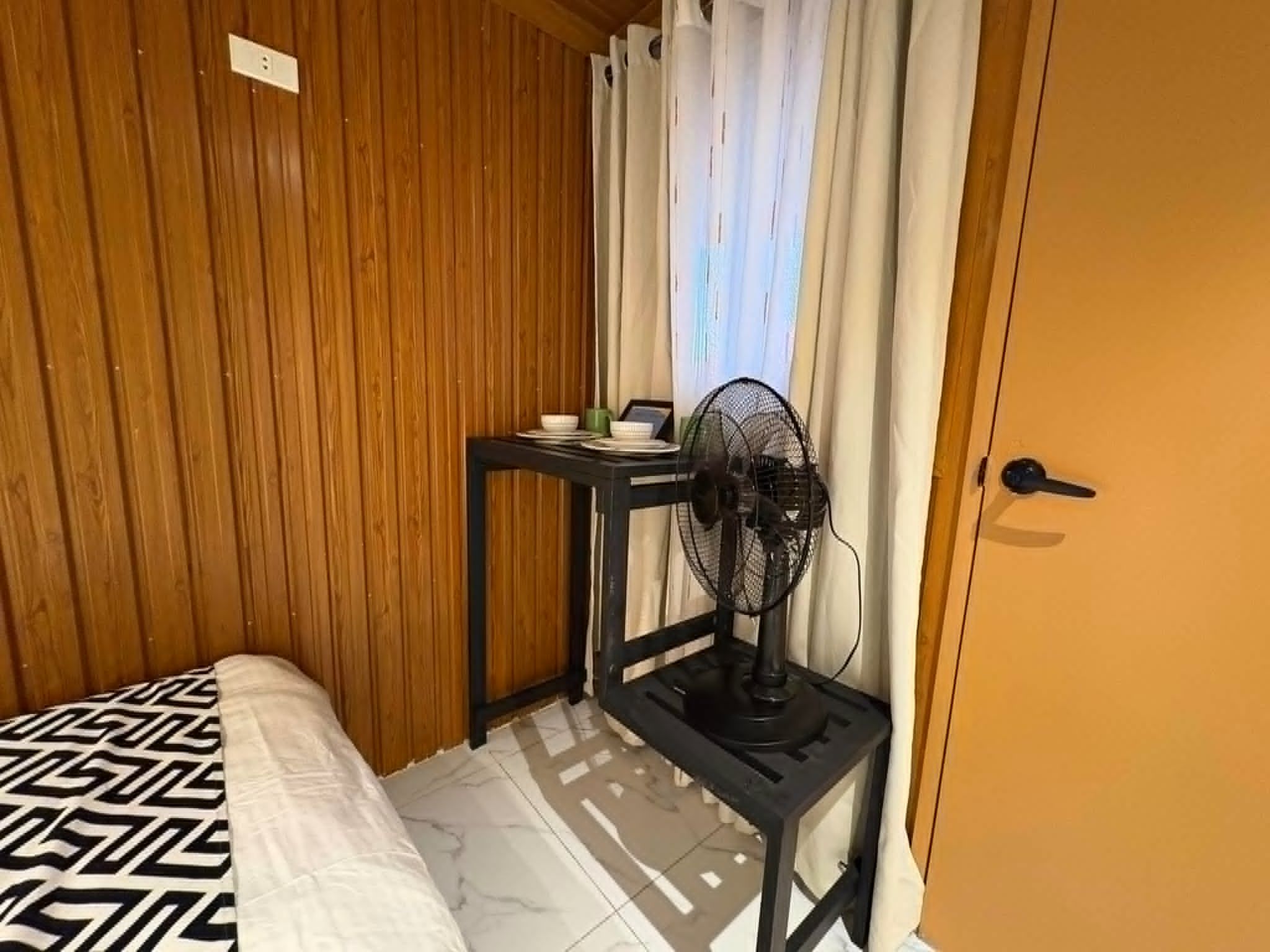 A small room with a bed, a table with a fan, and a door.