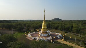 Un grand stupa doré, entouré d’une clôture blanche, avec des marches menant à son sommet, se dresse au milieu d’un paysage verdoyant et luxuriant, parsemé d’arbres et dominé par une colline lointaine.