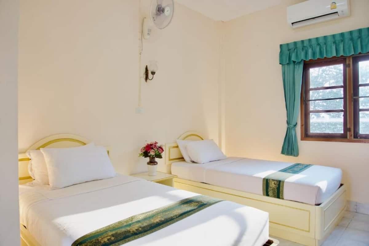 A hotel room with two single beds, a small table with a vase of flowers, a wall-mounted fan, an air conditioning unit, and a window with a green curtain.