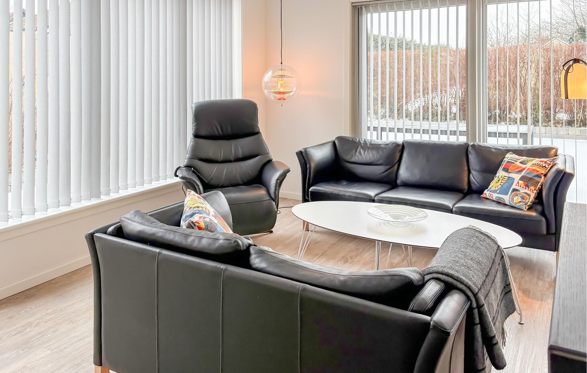 Een moderne woonkamer met zwarte lederen meubels, een witte salontafel en uitzicht door grote ramen met zonwering.