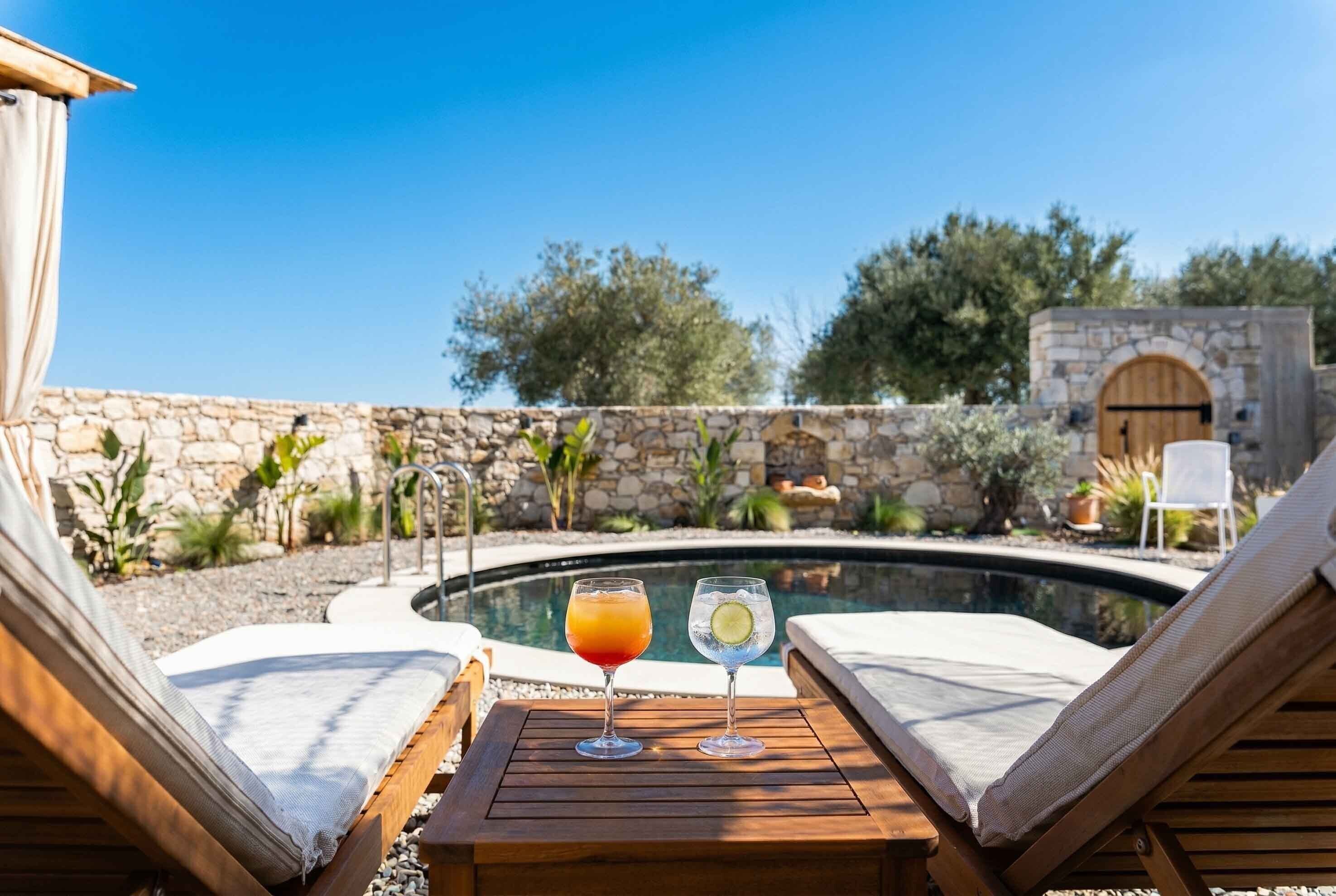A poolside lounge area with two lounge chairs, a wooden table, and two drinks.