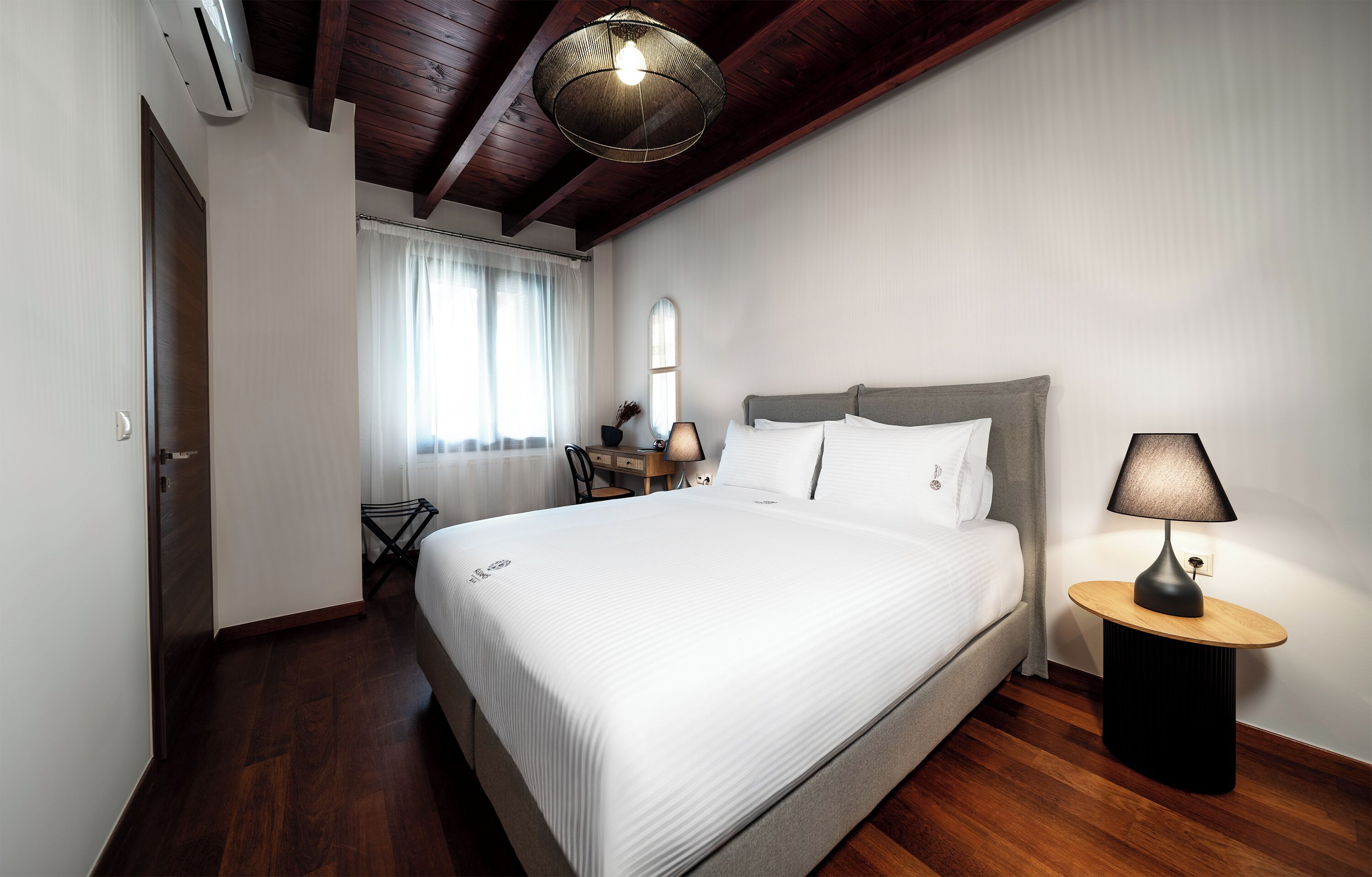 Una camera d'albergo con un letto grande, un comodino con una lampada, una finestra con tende e una ventola a soffitto.