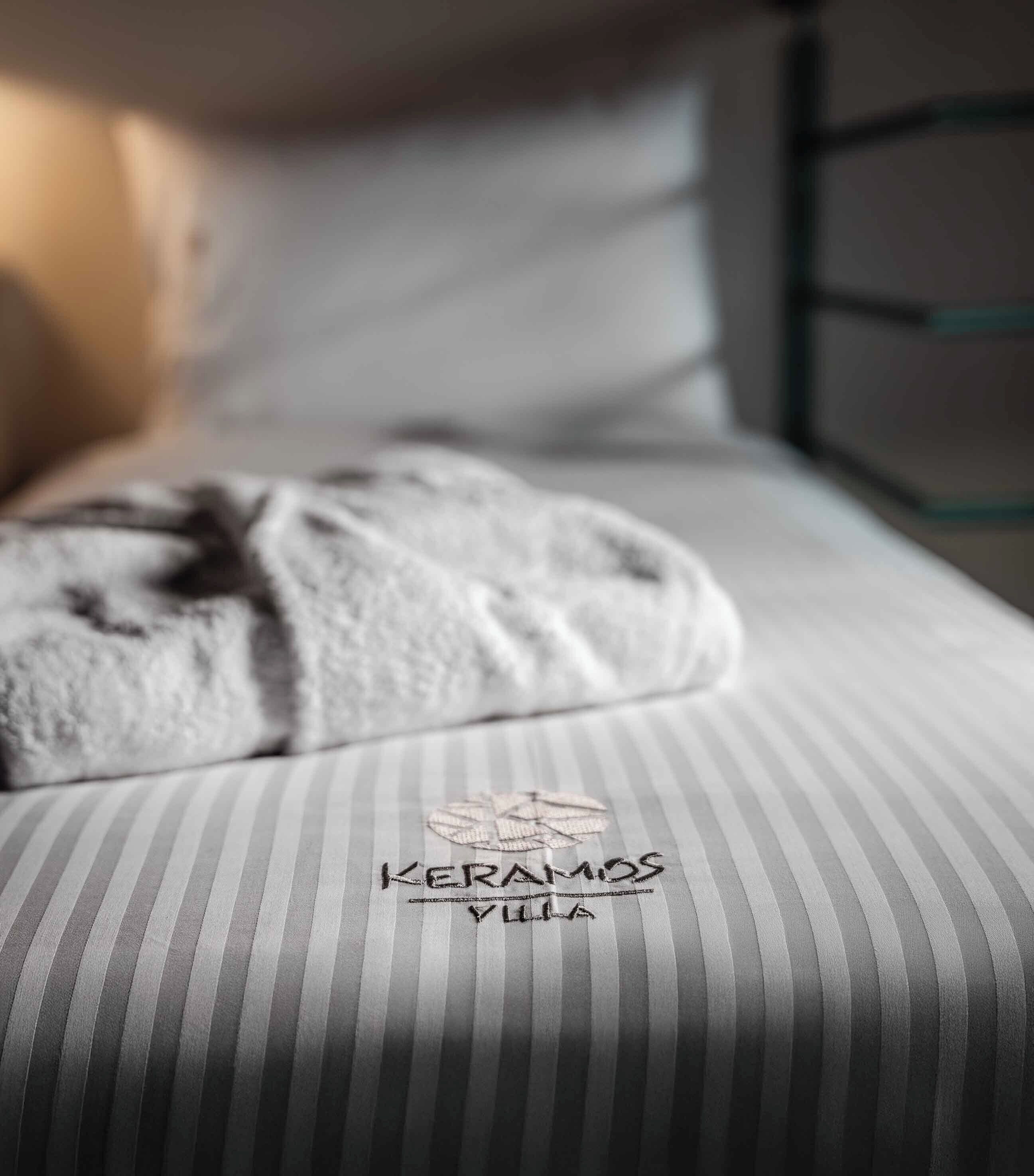 Una cama con sábana a rayas y una toalla. En la sábana se bordea el texto 'KERAMOS VILLA'.