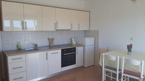 Een kleine keuken met witte kastjes, een vaatwasser en een kleine koelkast. Er is een eettafel met stoelen en een bloemenvaas.