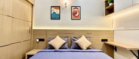 Un dormitorio moderno con una cama, dos almohadas, una mesita de noche, una lámpara de pared y dos cuadros enmarcados.