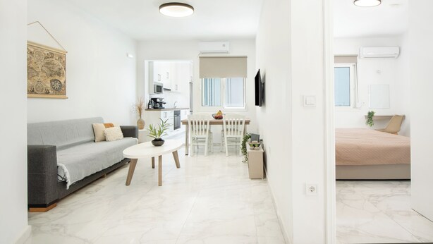 Ein modernes Wohnzimmer mit grauem Sofa, einem weißen Couchtisch und einem Essbereich mit weißem Tisch und Stühlen. Im Hintergrund befindet sich eine Küche mit weißen Schränken und einer Mikrowelle.