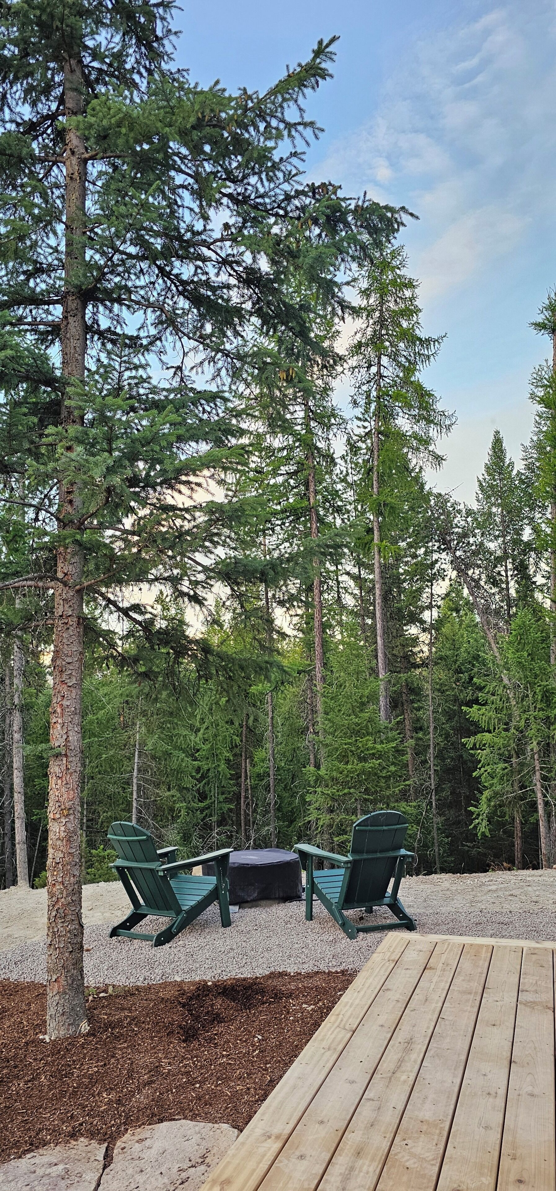 Een terras met twee groene Adirondack-stoelen, omgeven door een bos met hoge bomen.