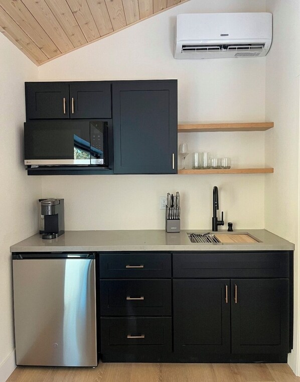 A modern kitchen with black cabinets, a stainless steel refrigerator, a microwave, and a coffee maker. There is a wooden shelf above the sink and a window above the microwave.