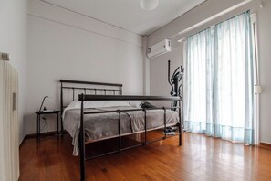 A bedroom with a metal bed frame, a bedside table, a wall-mounted air conditioner, and a window with sheer curtains. - Athenian Central Escape (Athens)