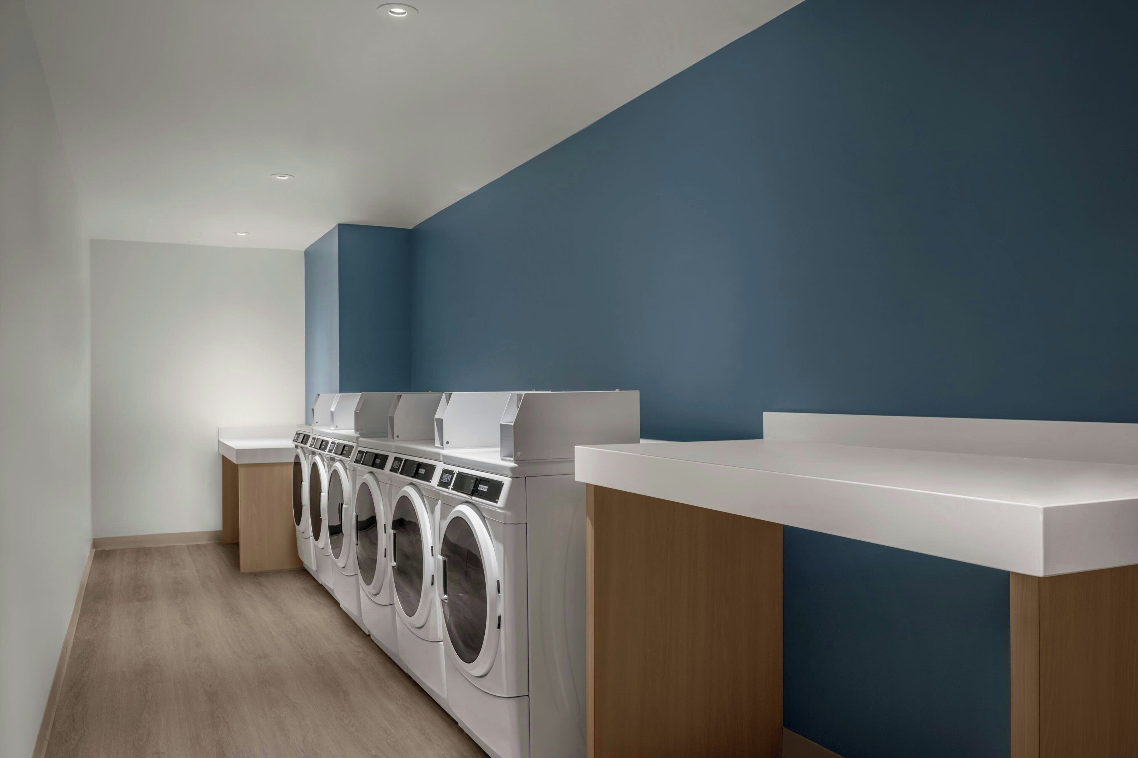 A laundry room with a row of front-loading washers and dryers, wooden cabinets, and a blue accent wall.