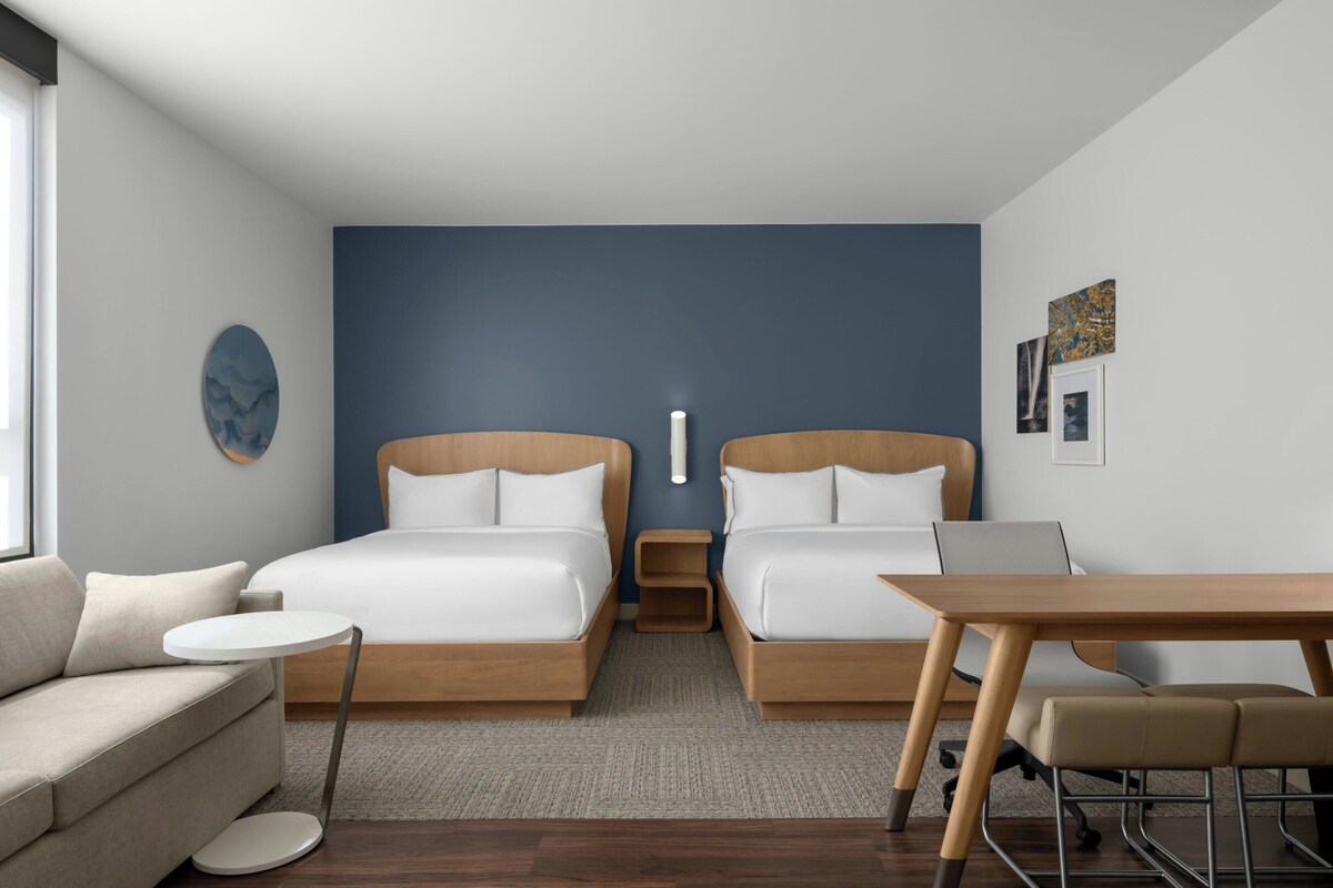 A modern hotel room with two beds, a sofa, a small table, and a desk.