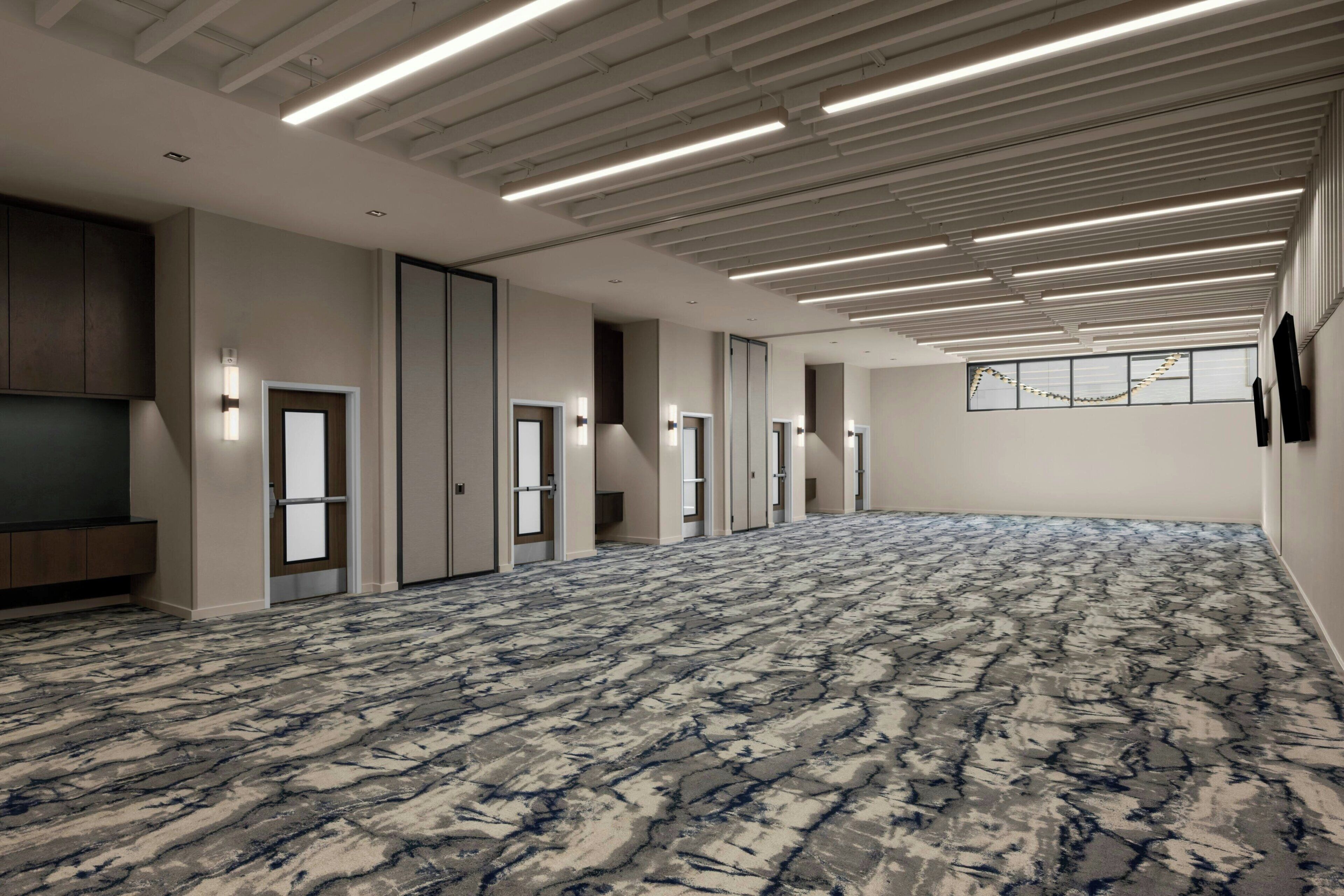 A spacious hotel room with a patterned carpet, multiple doors, and a ceiling with linear lighting fixtures.