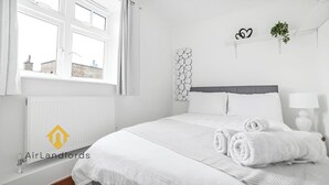 Ein Schlafzimmer mit Bett, weißer Bettwäsche, einem Heizkörper, einem Fenster mit Vorhängen, einer Wandpflanze und einem Logo.