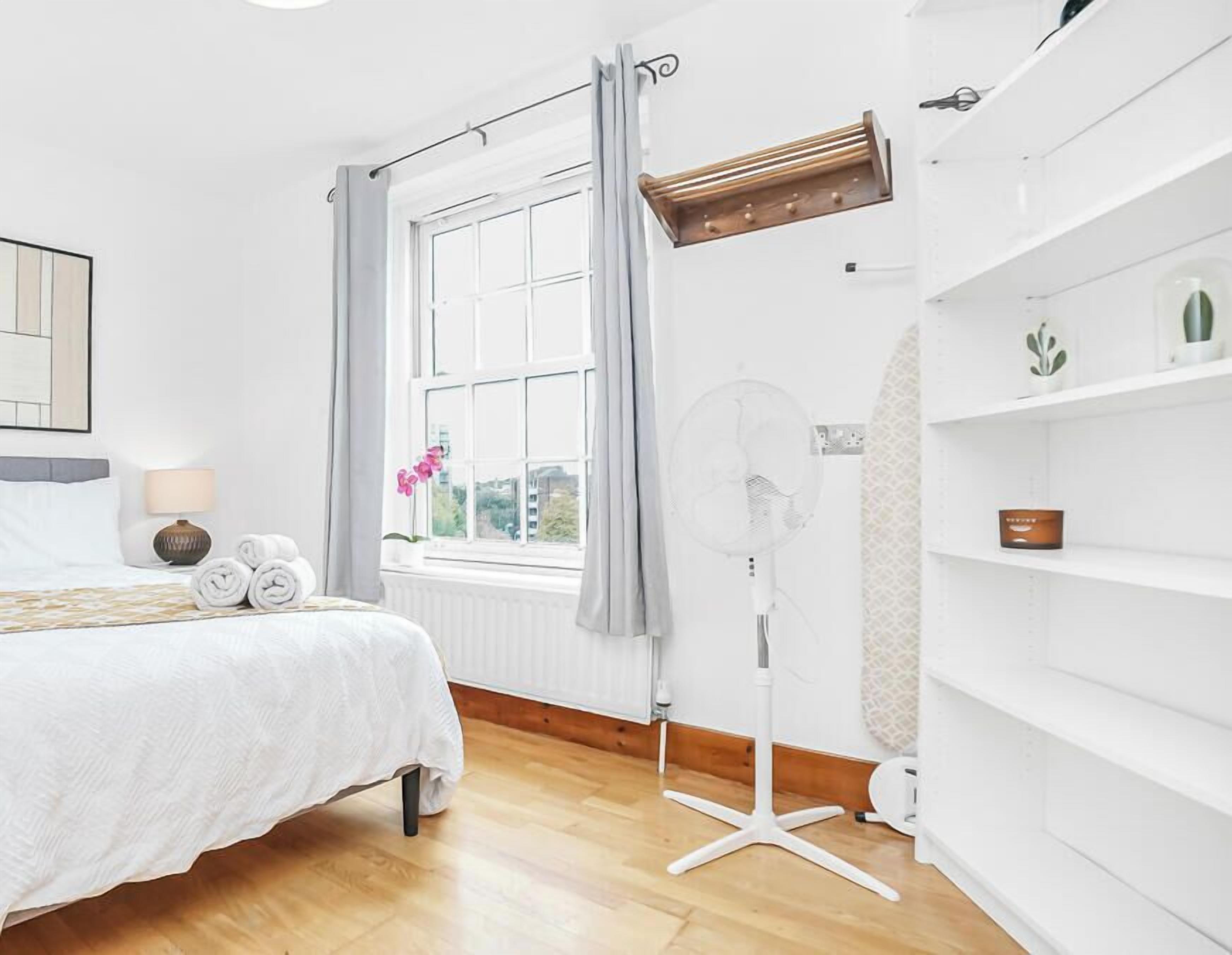 Ein Hotelzimmer mit Bett, Standventilator, einem fenster mit Vorhängen, einem Heizkörper und einem Regal mit einer Pflanze.