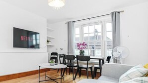 Ein modernes Wohnzimmer mit Flachbildfernseher, Essbereich mit Stühlen, einer Couch und einem großen Fenster mit Vorhängen.