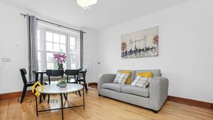 Ein modernes Wohnzimmer mit einem grauen Sofa, einem runden Tisch und zwei Stühlen. Es gibt ein Fenster mit Vorhängen, ein Gemälde an der Wand und eine Chandelier.