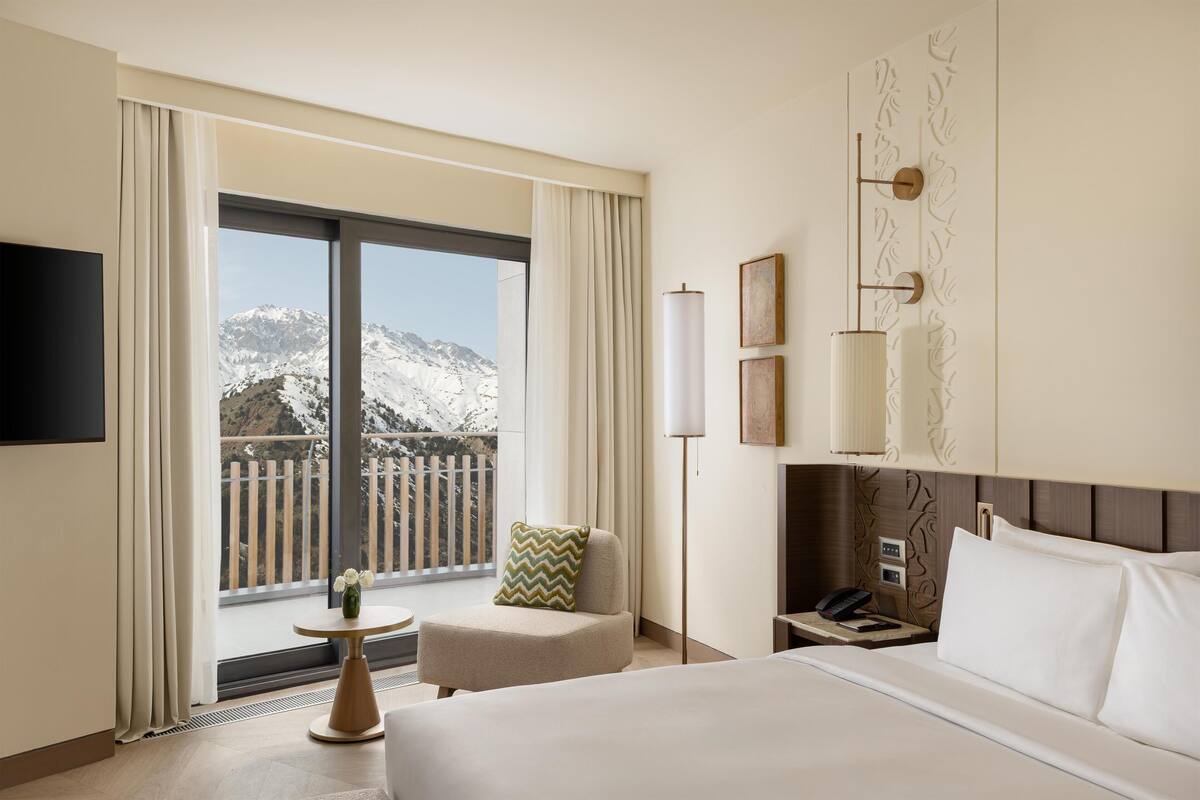 A hotel room with a bed, a chair, a bedside table with a lamp, a flat-screen TV, and a view of mountains.
