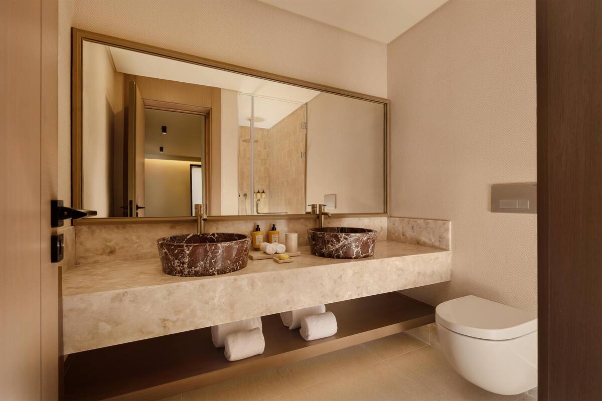 A modern bathroom with a large mirror, two marble sinks, a toilet, and a towel rack.
