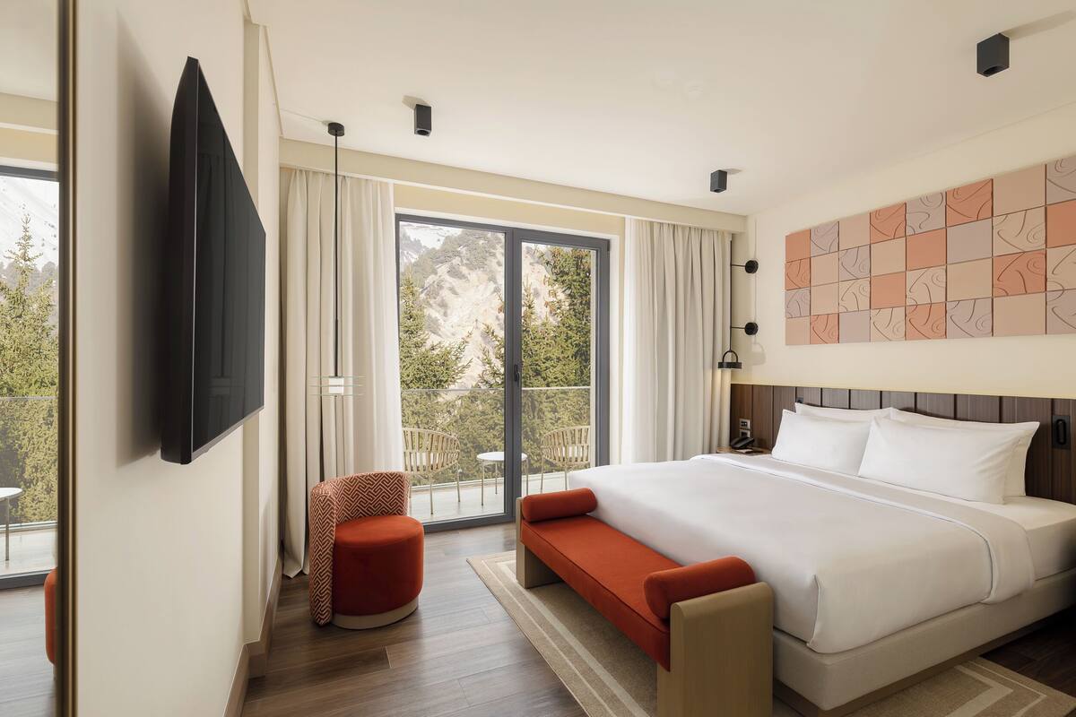 A modern hotel room with a large bed, a TV mounted on the wall, a balcony with outdoor seating, and a view of the outdoors.