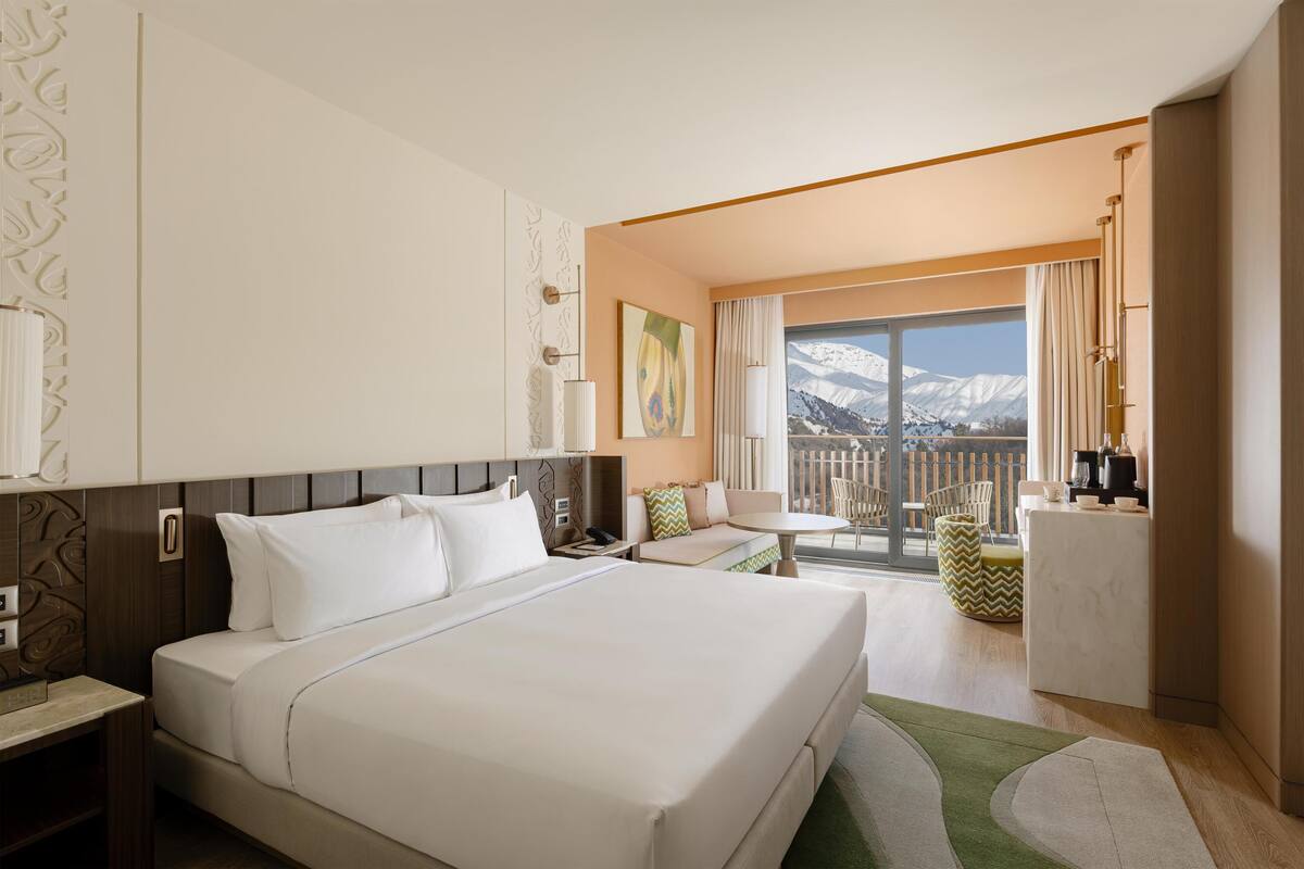 A hotel room with a large bed, a desk, a chair, and a balcony with a mountain view.