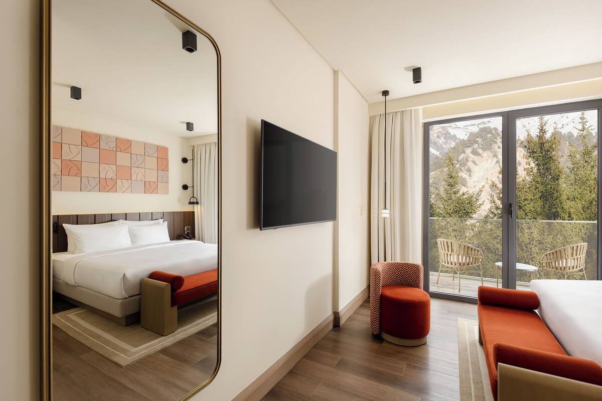 Una habitación de hotel moderna con cama, televisor, balcón con sillas y un espejo grande.