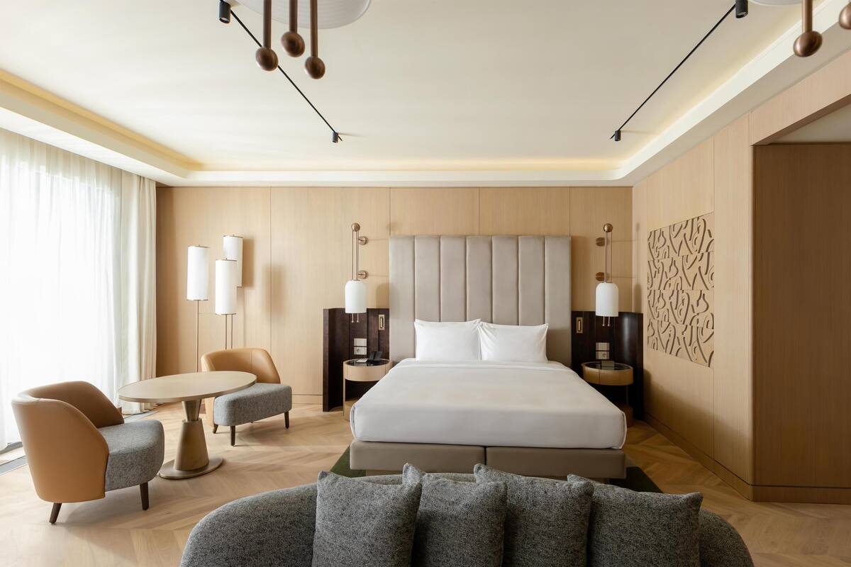 A modern hotel room with a large bed, two bedside tables, a sofa, a small round table, and a decorative wall panel.