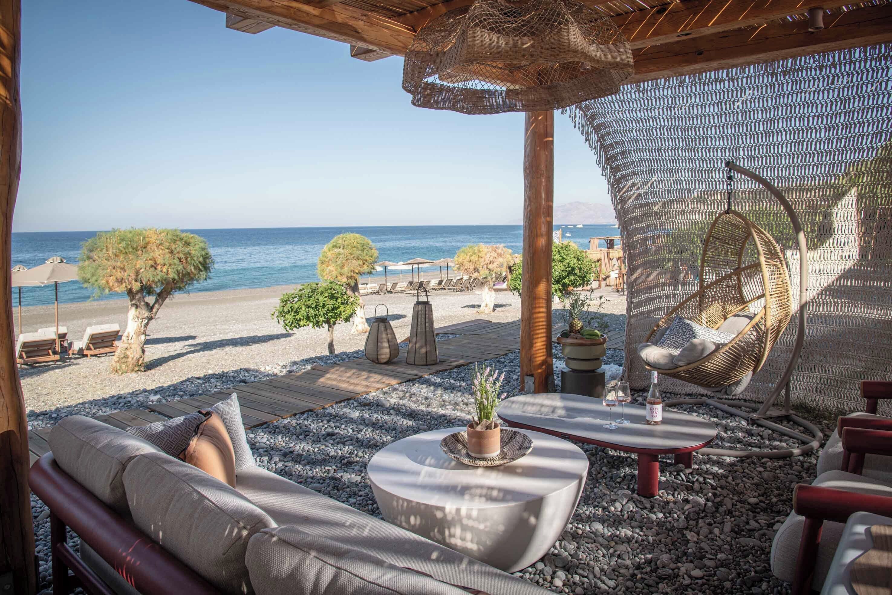 A beachside lounge with a view of the sea, featuring a wicker chair, a sofa, and a small table with a potted plant.