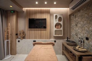 Ein moderner Wellnessraum mit Massagetisch, Holzvertäfelung, einem Fernseher und einem an der Wand befestigten Ablagebereich mit Handtüchern.