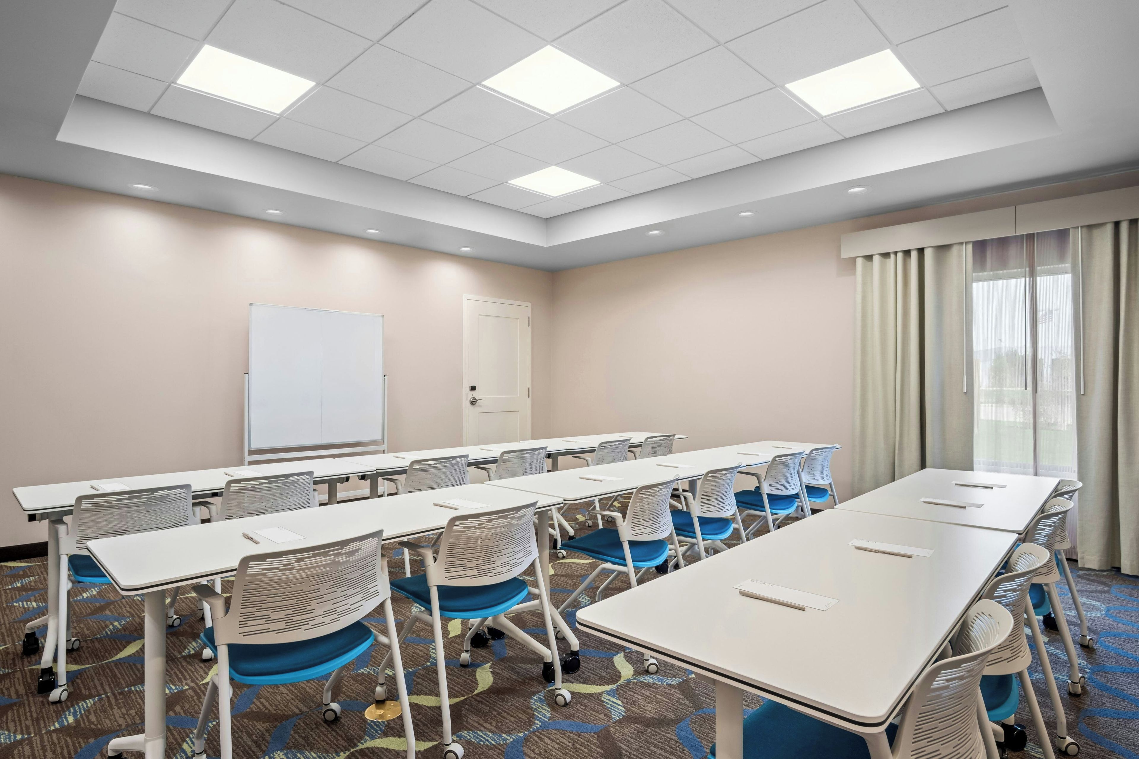 Una sala de conferencias con mesas blancas, sillas azules, una pizarra y una puerta.