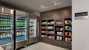 Interior de una tienda de comestibles con dispensadores refrigerados para bebidas, estantes con aperitivos y un letrero que dice 'The Market'.