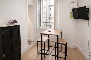 Ein kleines Zimmer mit Holzboden, einem schwarzen Metalltisch mit Stuhl, einem Schrank, einem Fenster mit Blick auf Gebäude und einem an der Wand befestigten Fernseher.