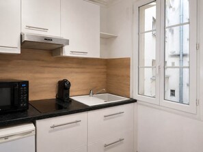 A modern kitchen with white cabinets, a black countertop, a built-in microwave, a coffee maker, and a window with a view of buildings. - Studio Parisien grands boulevards Couple ou Solo (Paris)