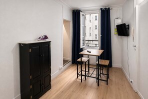 Ein kleines Zimmer mit Holzboden, einem schwarzen Schrank, einem Tisch mit zwei Stühlen, einem Fenster mit blauen Vorhängen und einem an der Wand montierten Fernseher.