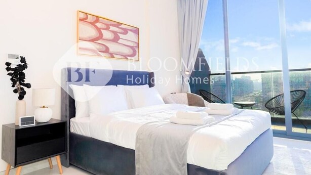 A modern bedroom with a large bed, a nightstand, a lamp, and a view of the city through large windows. - Bloomfields CharmingStu Samana view (Dubai)