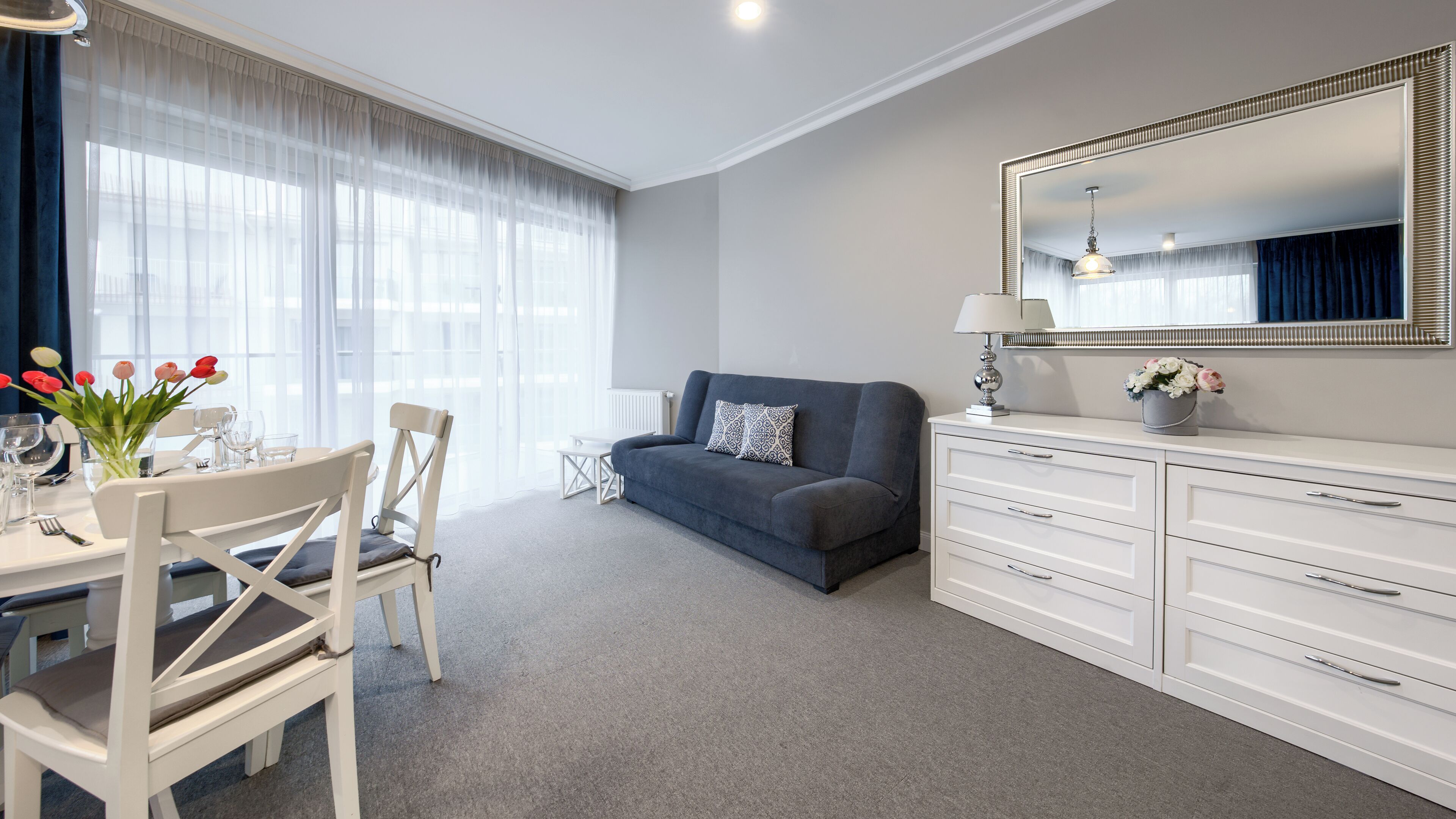 A modern hotel room with a grey sofa, a dining table set with glasses and cutlery, a white dresser, and a large mirror.