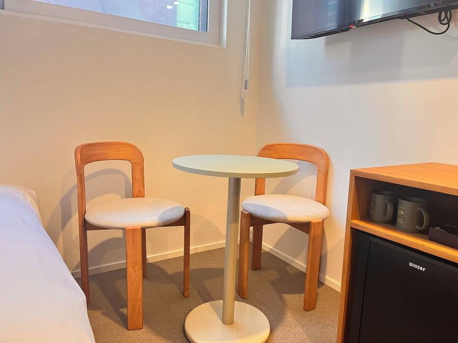 A small room with two wooden chairs, a round table, a bed, a TV mounted on the wall, and a shelf with mugs.